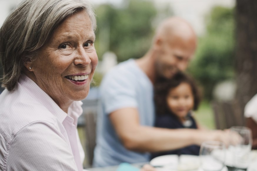 Eldre dame sitter i hagen rundt spisebord med familie og ser i kamera